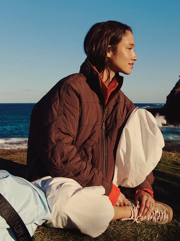 Free People Movement Pippa Packable Puffer in Cocoa worn with white pants while seated by the ocean | Twentyseven Toronto