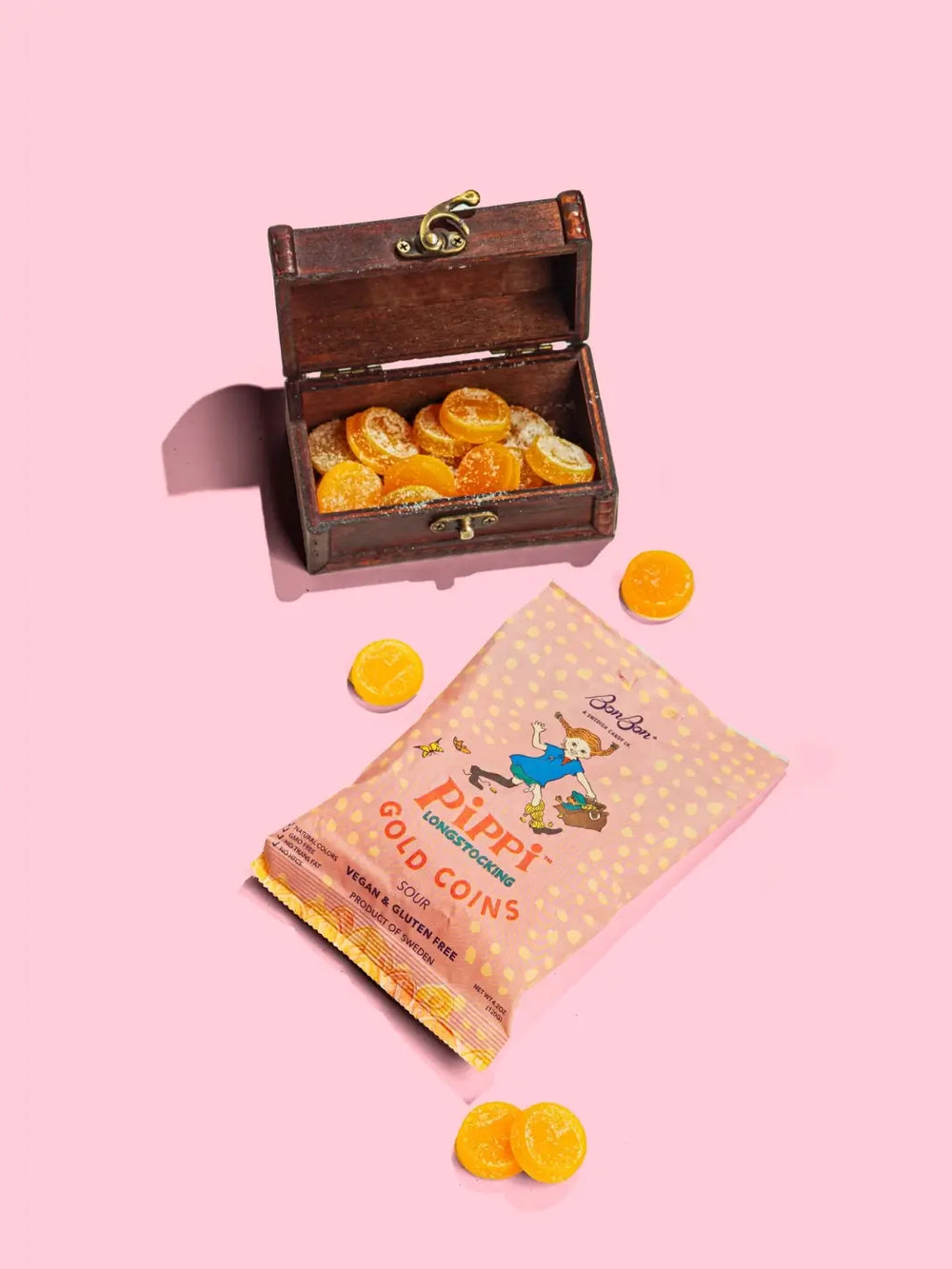 A small, open wooden treasure chest is filled with sugary yellow treats next to a pink bag of Pippi Longstocking Gold Coins Swedish Candy from BonBon NYC on a bright pink background | Twentyseven Toronto