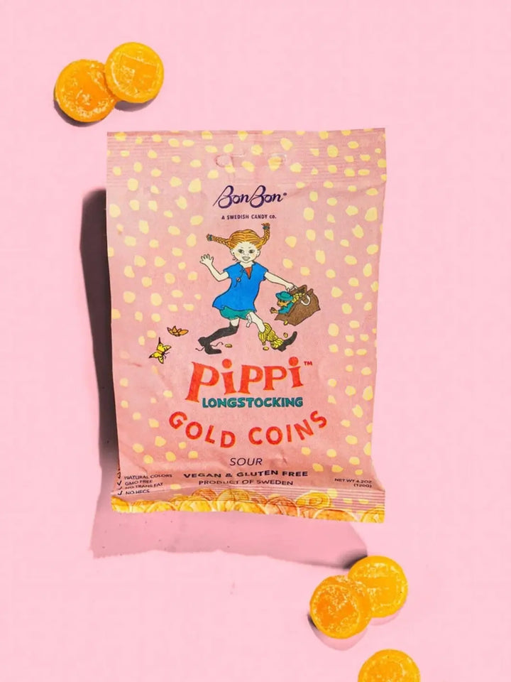 A pink pouch of Pippi Longstocking Gold Coins Swedish Candy from BonBon NYC sits on a pink backdrop, surrounded by a few loose, round sugar-dusted yellow candy | Twentyseven Toronto
