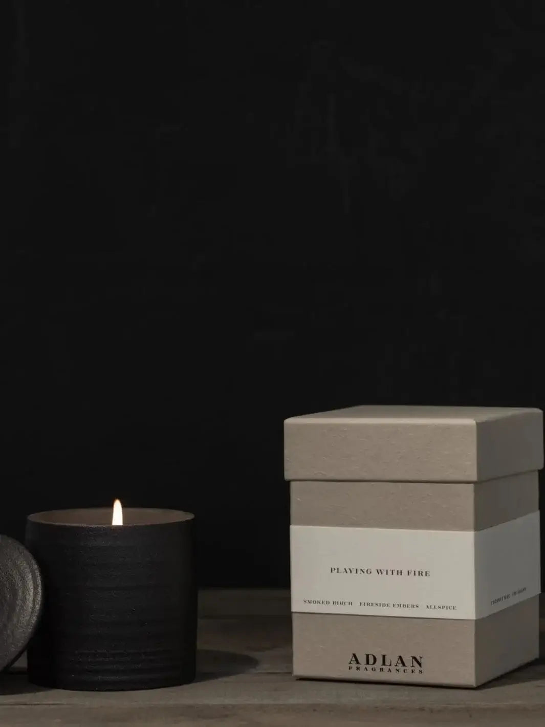 A lit Adlan Fragrances Playing with Fire in Petite candle glows in its black textured vessel next to its matching grey product packaging box against a dark background | Twentyseven Toronto