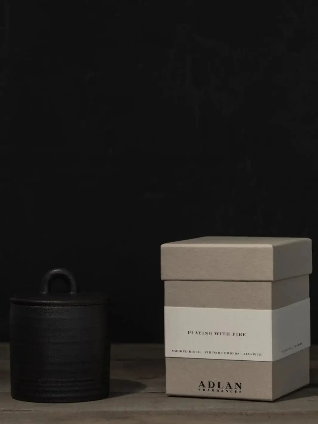 The Adlan Fragrances Playing with Fire in Petite candle is shown enclosed with its black lid on, beside its minimalist grey branding box on a wooden surface | Twentyseven Toronto