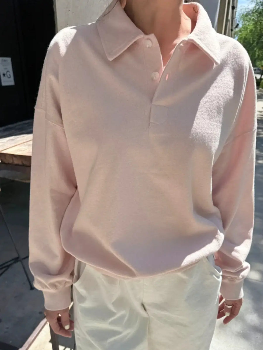 A person wears a light pastel pink Le Bon Shoppe Polo Sweatshirt in Baby Pink with a partially buttoned collar and white pants standing outdoors | Twentyseven Toronto