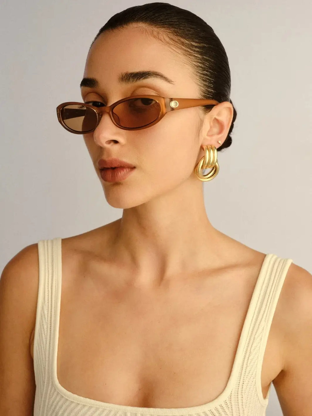 A woman wearing oval-framed Le Specs Popalicious + | Pearl Caramel sunglasses paired with thick gold hoop earrings | Twentyseven Toronto
