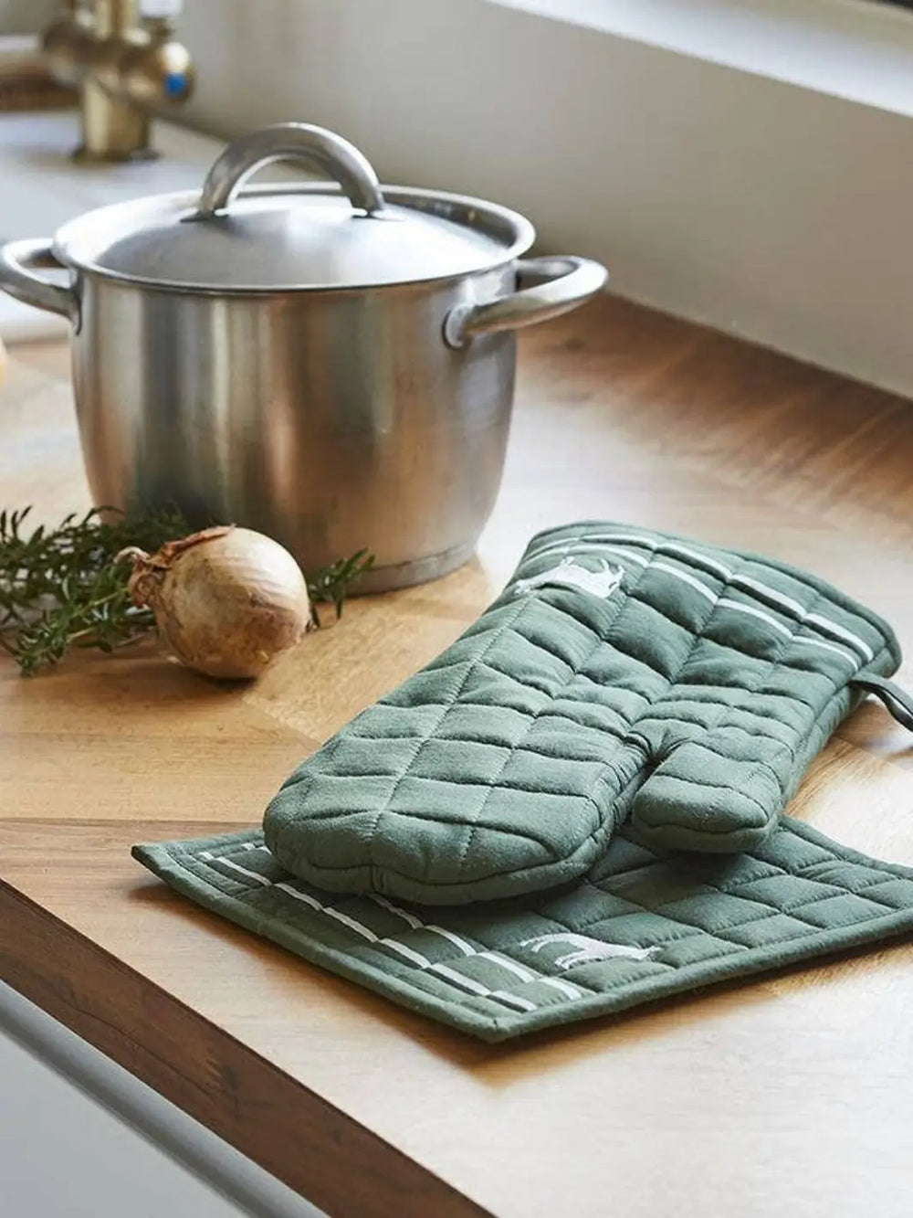 Olive green pot holder and trivet set from Sylvie Thiriez on a wooden kitchen counter with a steel pot and onion | Twentyseven Toronot