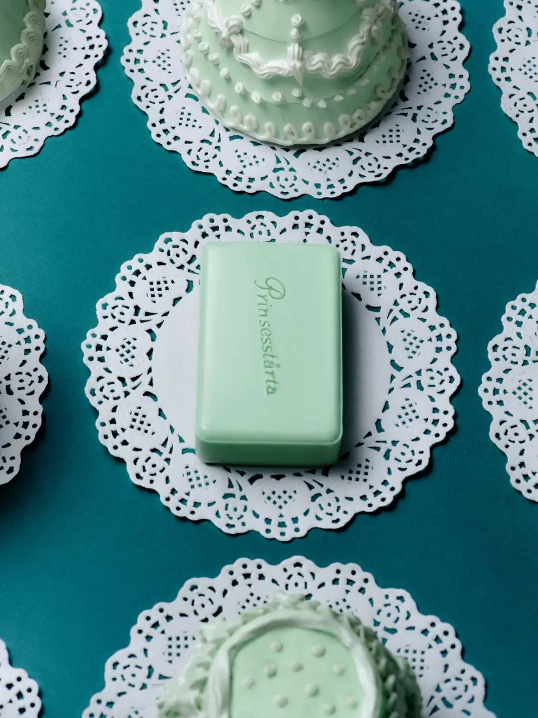 Top-down view of the mint green Flamingo Estate Prinsesstarta Milk Bar soap rest on a white lace paper doily surrounded by decorative mini cakes | Twentyseven