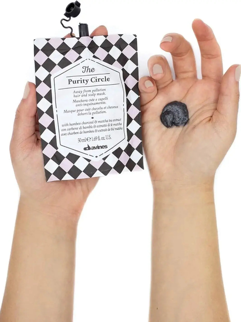 A pair of hands holds a grey and black checkered pouch of Davines The Purity Circle of Chronicles Hair Mask next to an open palm containing a dollop of dark grey product against a white background | Twentyseven Toronto