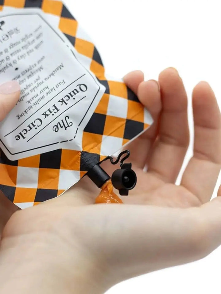 A pair of hands squeezes a dollop of orange cream product from an orange and black checkered pouch of Davines The Quick Fix Circle of Chronicles Hair Mask into an open palm against a white background | Twentyseven Toronto