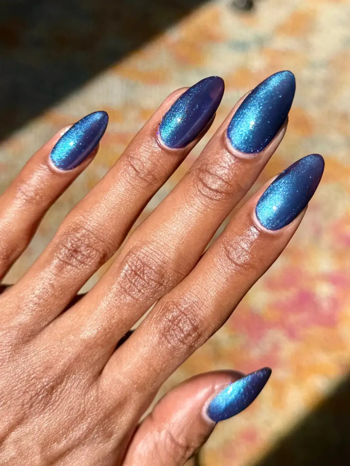 A close-up view shows a hand over a patterned rug, highlighting the dark violet base and bright blue magnetic line of Death Valley Nails Quietude (Or Swallowing Certain Unhappiness) Magnetic Nail Polish | Twentyseven Toronto