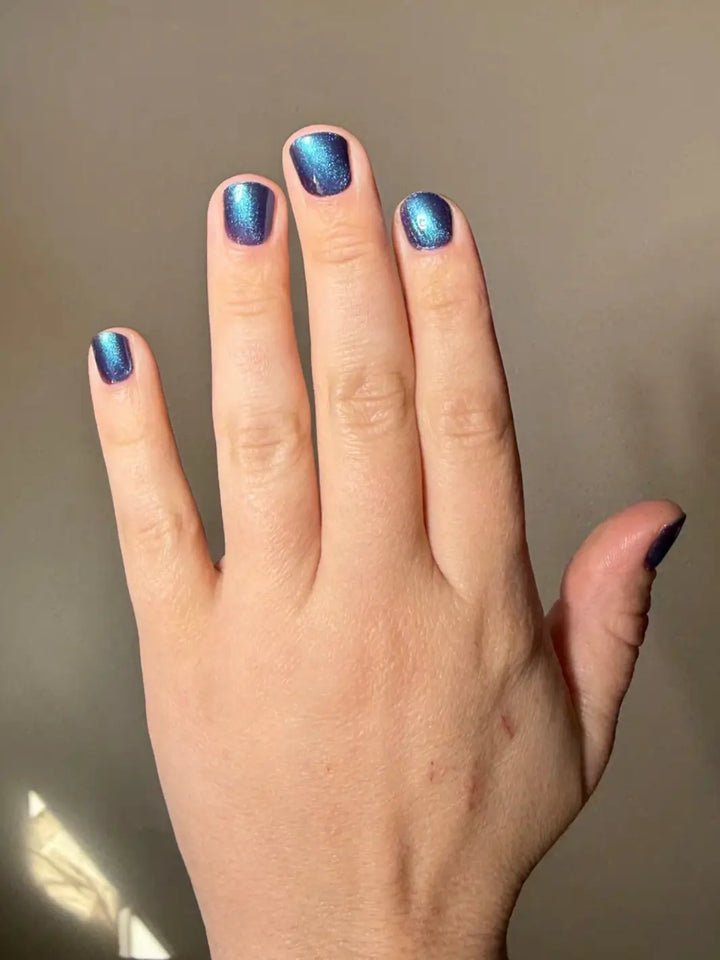 A hand with short, square nails displays a smooth, shimmering blue shade of Death Valley Nails Quietude (Or Swallowing Certain Unhappiness) Magnetic Nail Polish against a plain gray background | twentyseven Toronto