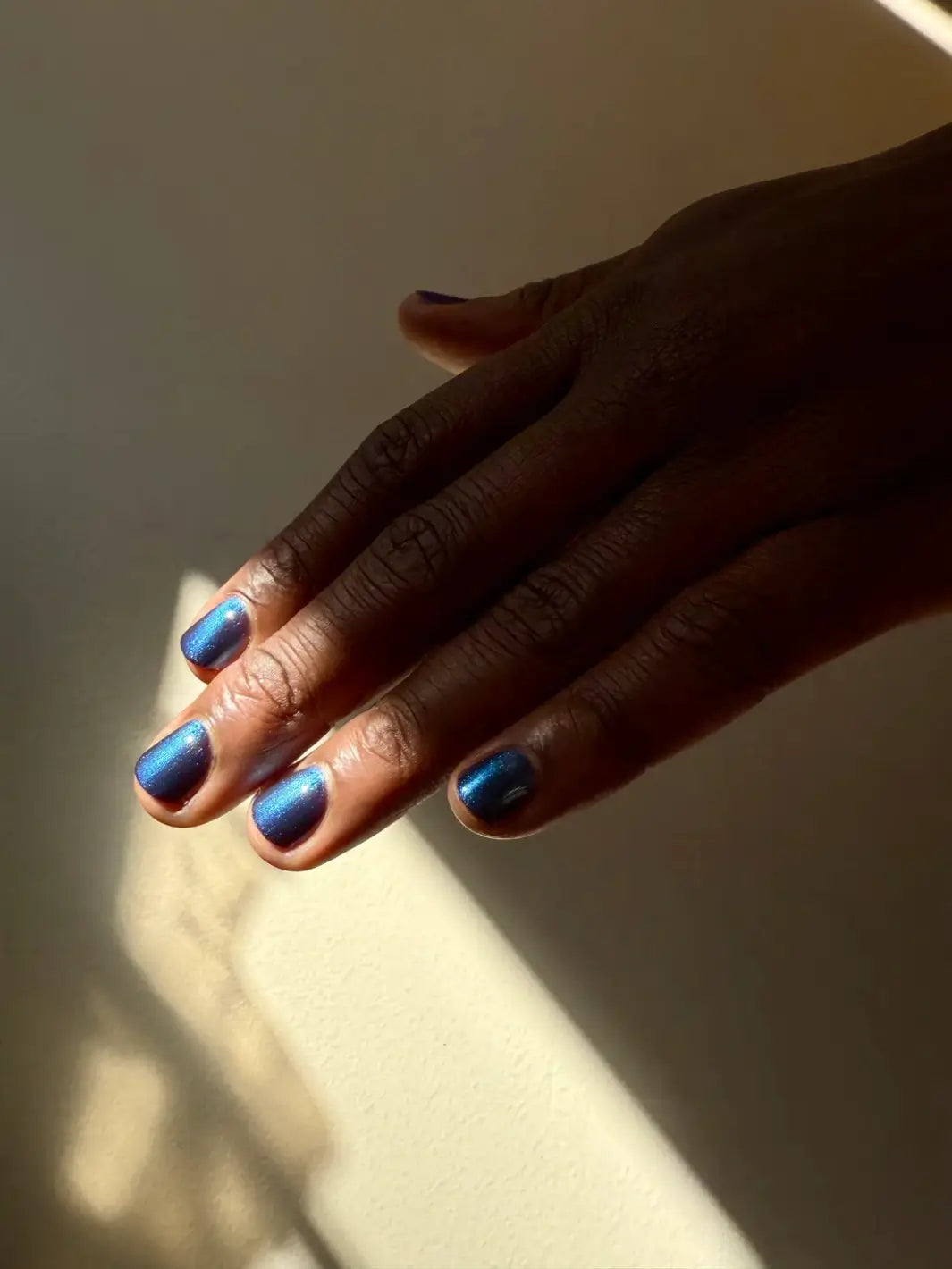 A hand with a deep skin tone and short nails is positioned in a stream of sunlight to reveal the dark blue tones of Death Valley Nails Quietude (Or Swallowing Certain Unhappiness) Magnetic Nail Polish | Twentyseven Toronto