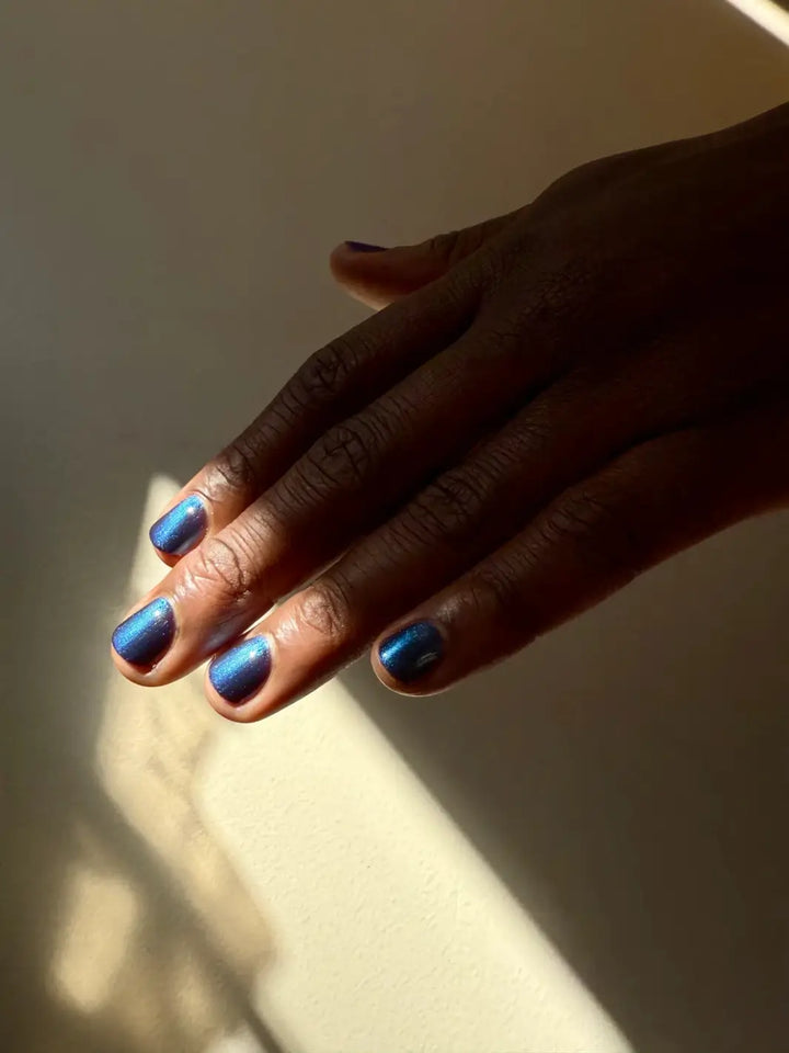 A hand with a deep skin tone and short nails is positioned in a stream of sunlight to reveal the dark blue tones of Death Valley Nails Quietude (Or Swallowing Certain Unhappiness) Magnetic Nail Polish | Twentyseven Toronto