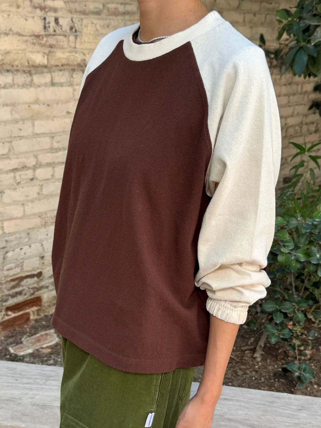 Side view of Heavy Naturelle Tee in Chestnut/Naturel featuring a chocolate brown body, cream raglan sleeves, crew neckline and relaxed fit, styled with olive green pants | Twentyseven Toronto