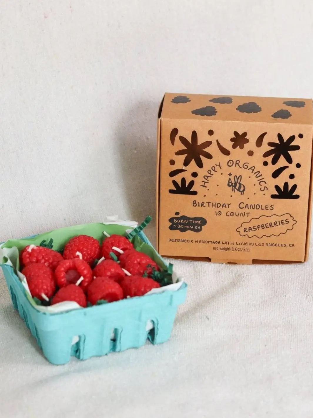 Carton of red birthday candles shaped like raspberries next to a box labeled 'Happy Organics' on a light background | Twentyseven Toronto