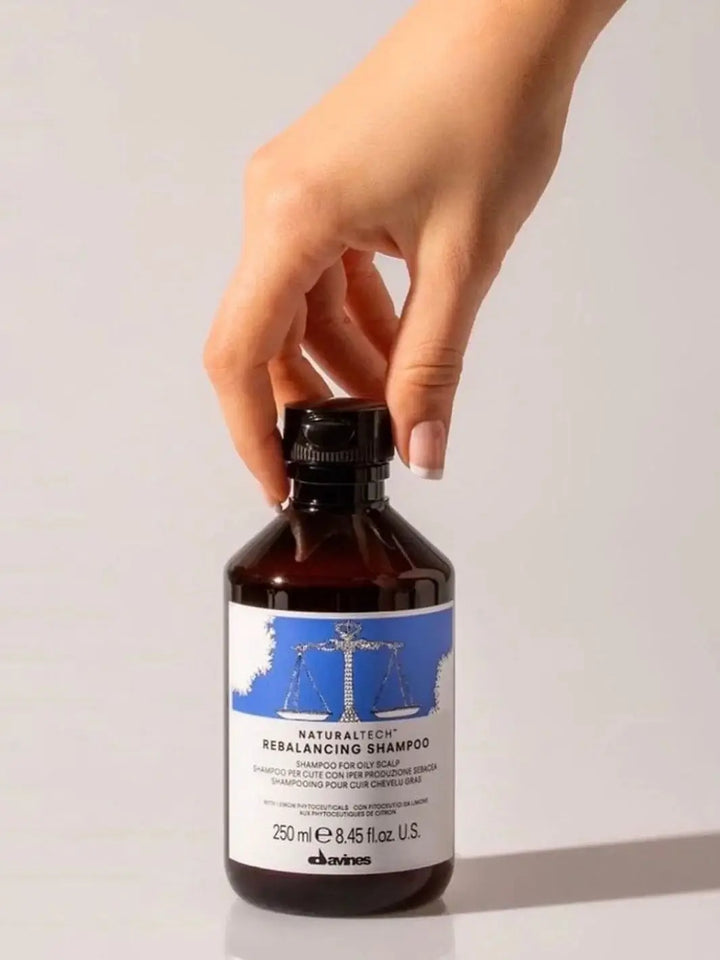 A hand reaches down to touch the black cap of a bottle of Davines REBALANCING Shampoo against a beige background | Twentyseven Toronto