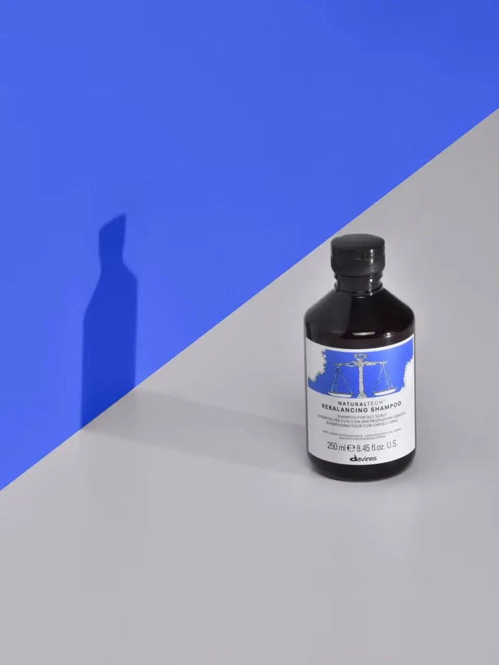 A bottle of Davines REBALANCING Shampoo on a light grey surface, casting a shadow against a bright blue wall | Twentyseven Toronto