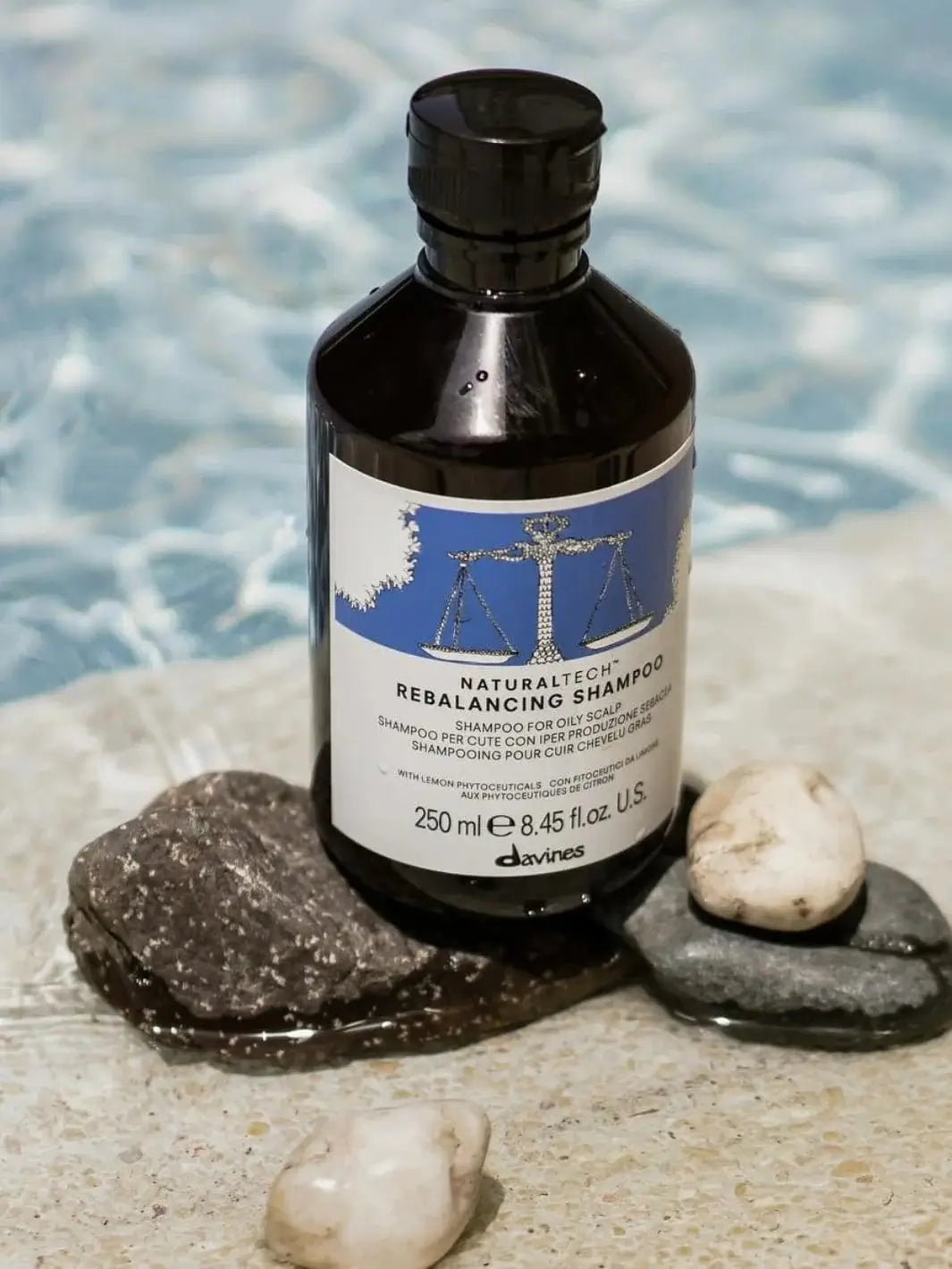 A bottle of Davines REBALANCING Shampoo is balanced on smooth river stones at the edge of a swimming pool with rippling blue water | Twentyseven Toronto