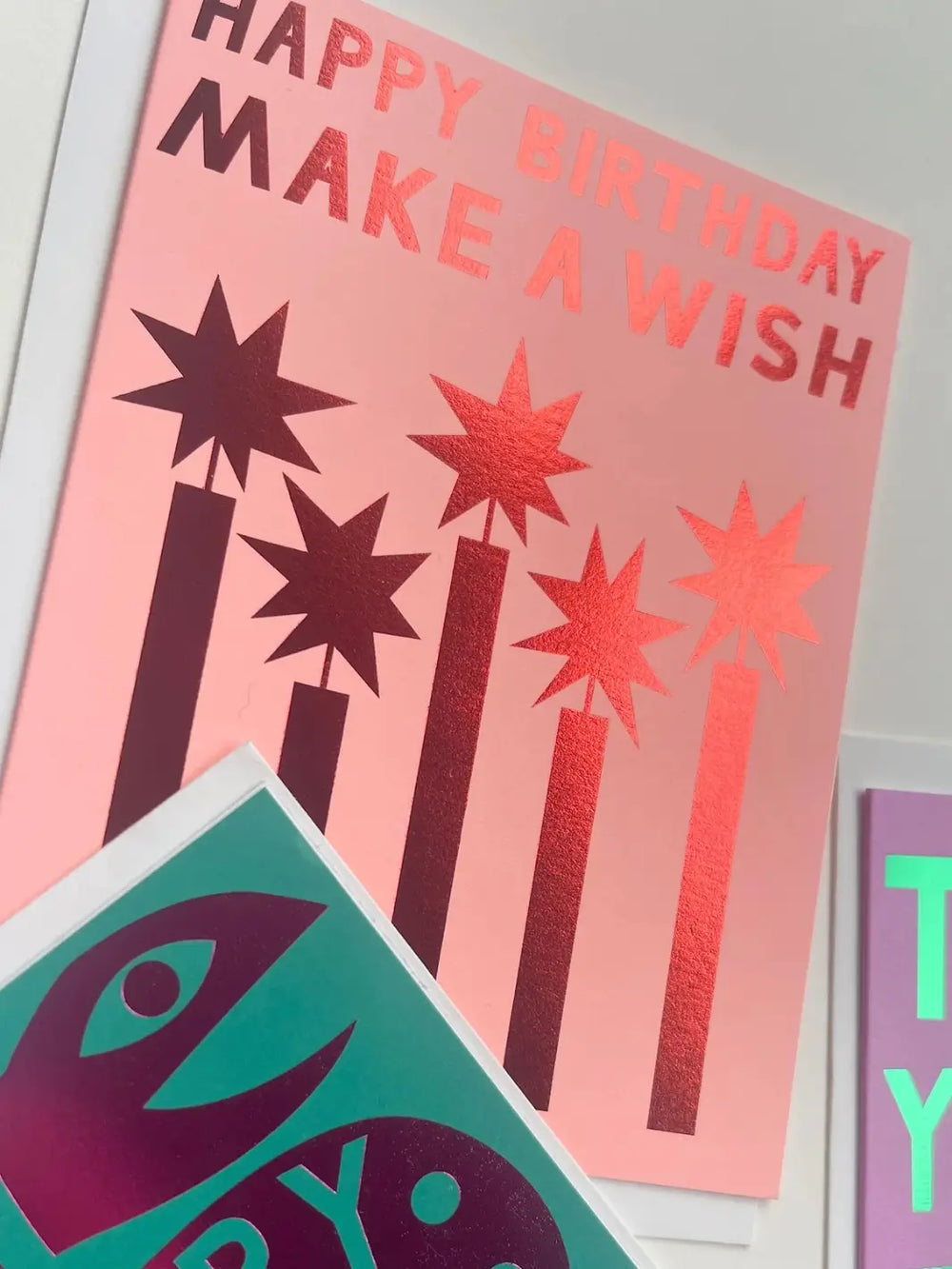 A close-up, angled view shows the pink Banquet Workshop Red Foil Happy Birthday Make a Wish Note Card featuring metallic red foil candles and text, partially layered behind a teal greeting card | Twentyseven