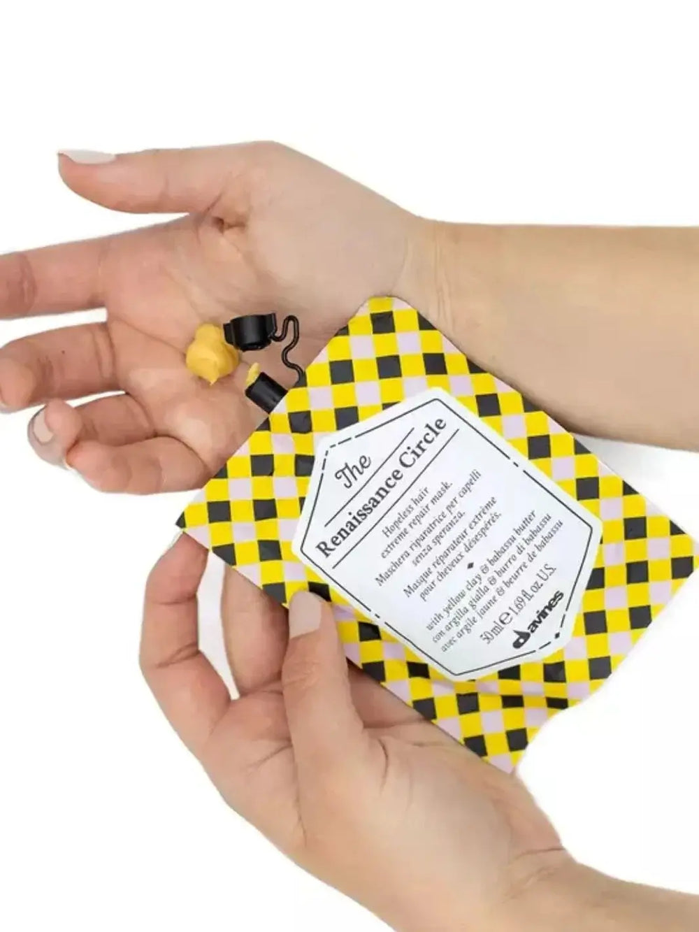 A pair of hands squeezes a small dollop of yellow cream product from a checkered pouch of Davines The Renaissance Circle of Chronicles Hair Mask into an open palm | Twentyseven Toronto