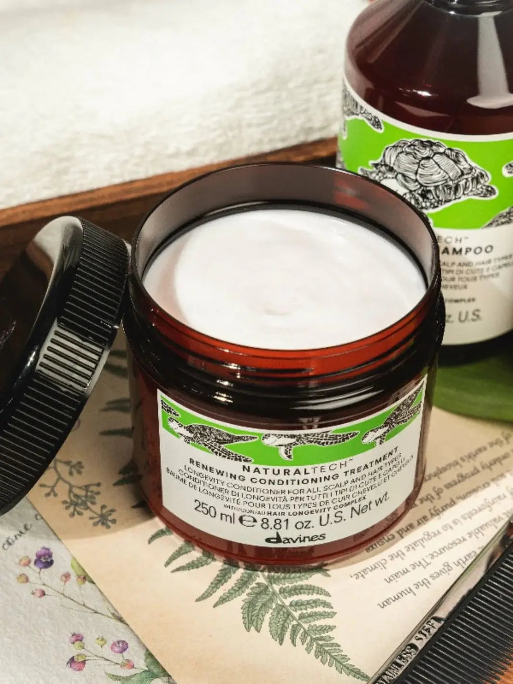 An open jar filled with smooth white cream showcases the Davines RENEWING Conditioning Treatment next to its black lid and a matching shampoo bottle on a decorated surface | Twentyseven Toronto