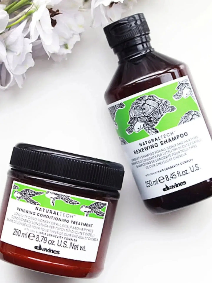 A bottle of Davines RENEWING Shampoo sits on a white surface next to a tub of the matching conditioning treatment and white flowers | Twentyseven Toronto