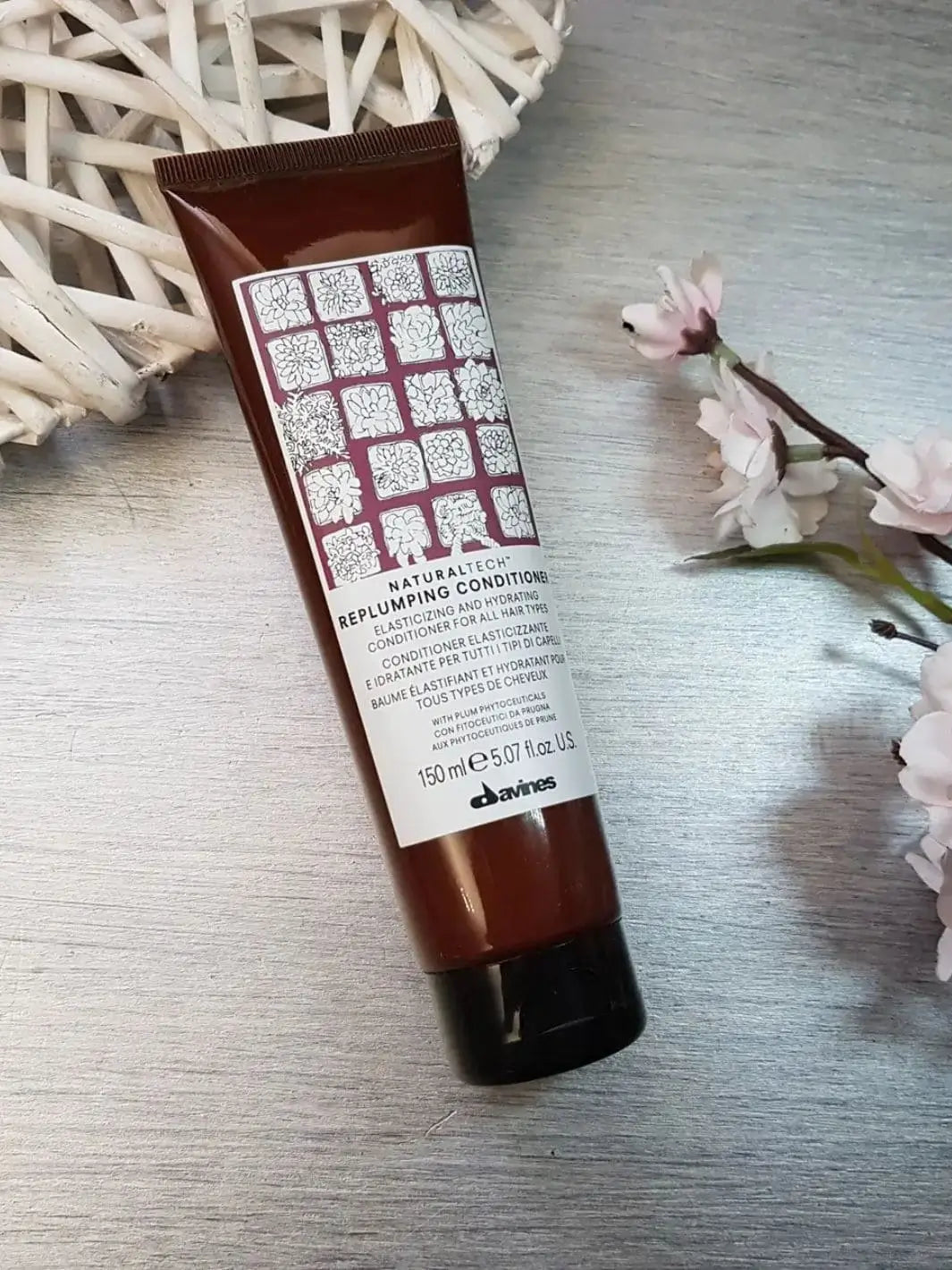 A single tube of Davines REPLUMPING Conditioner is placed on a wooden surface beside a white woven object and small pink flowers | Twentyseven Toronto