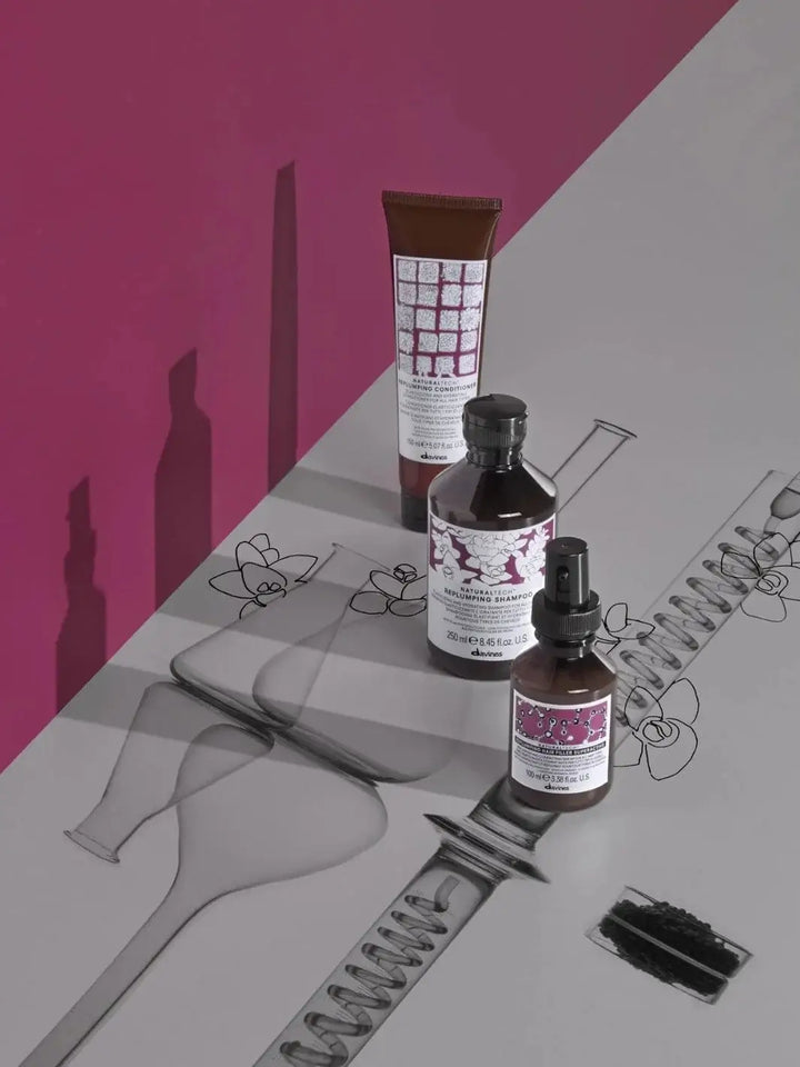 A tube of Davines REPLUMPING Conditioner stands in a line with a matching shampoo bottle and spray bottle against a split pink and grey background with illustrations | Twentyseven Toronto