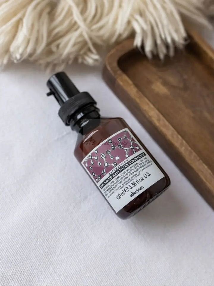 An amber spray bottle of Davines REPLUMPING Hair Filler Superactive rests on a white textured surface next to a small wooden tray | Twentyseven Toronto