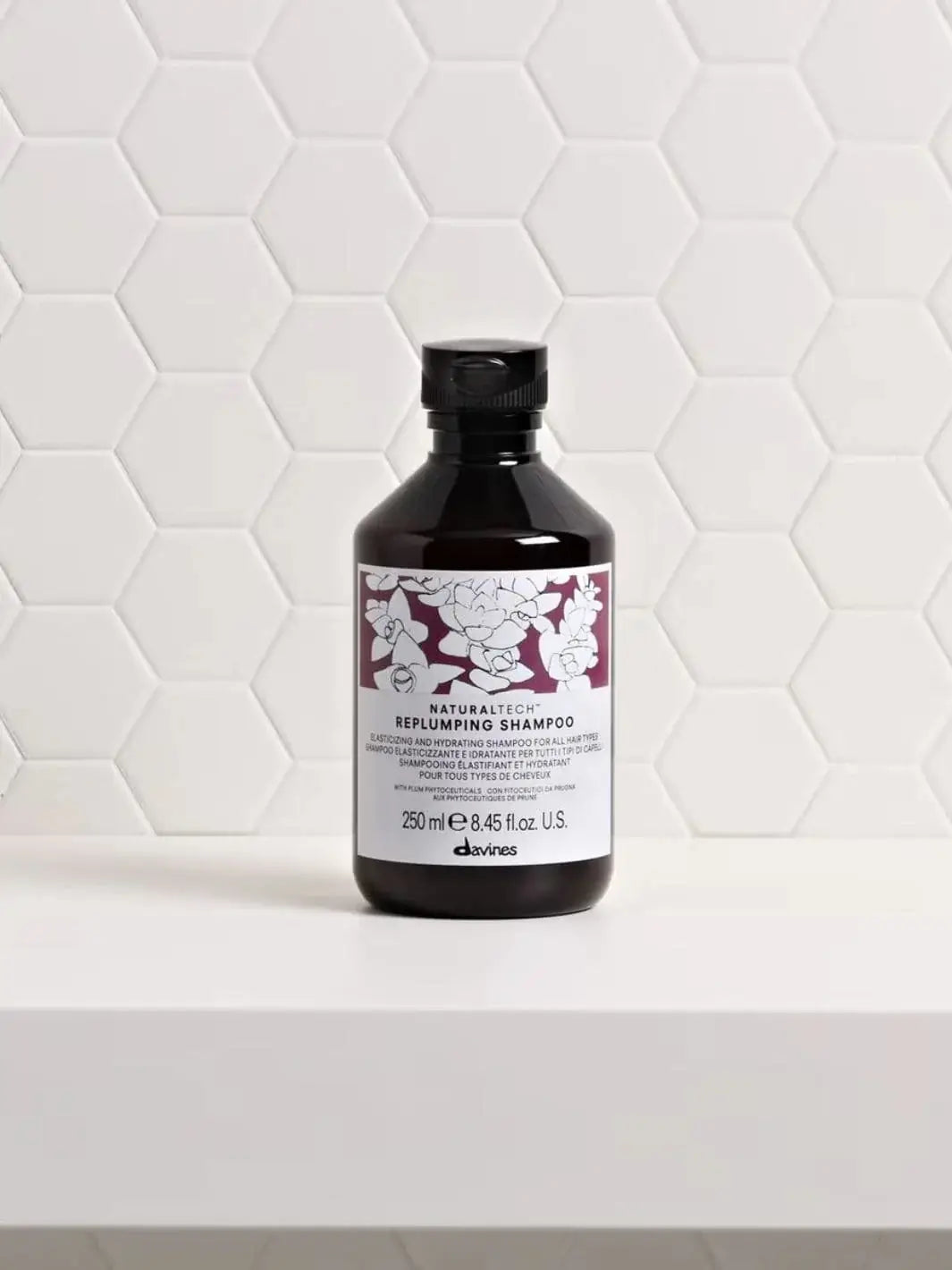 A bottle of Davines REPLUMPING Shampoo sits on a white shelf against a white tile background | Twentyseven Toronto