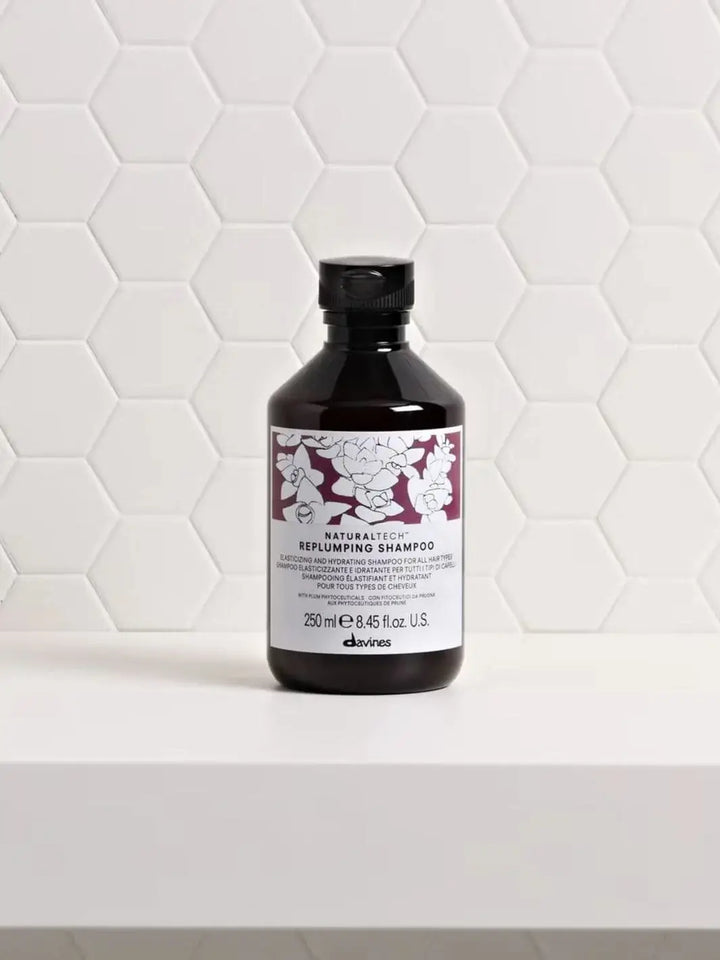 A bottle of Davines REPLUMPING Shampoo sits on a white shelf against a white tile background | Twentyseven Toronto