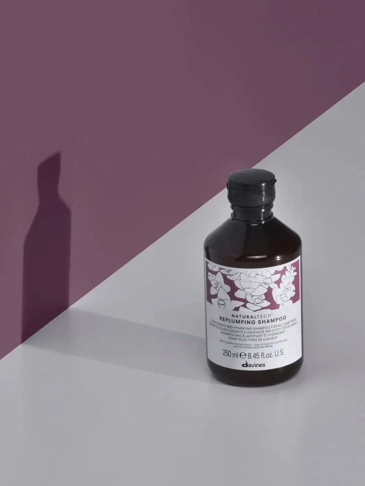 A bottle of Davines REPLUMPING Shampoo stands on a grey surface casting a shadow against a solid purple wall backdrop | Twentyseven Toronto