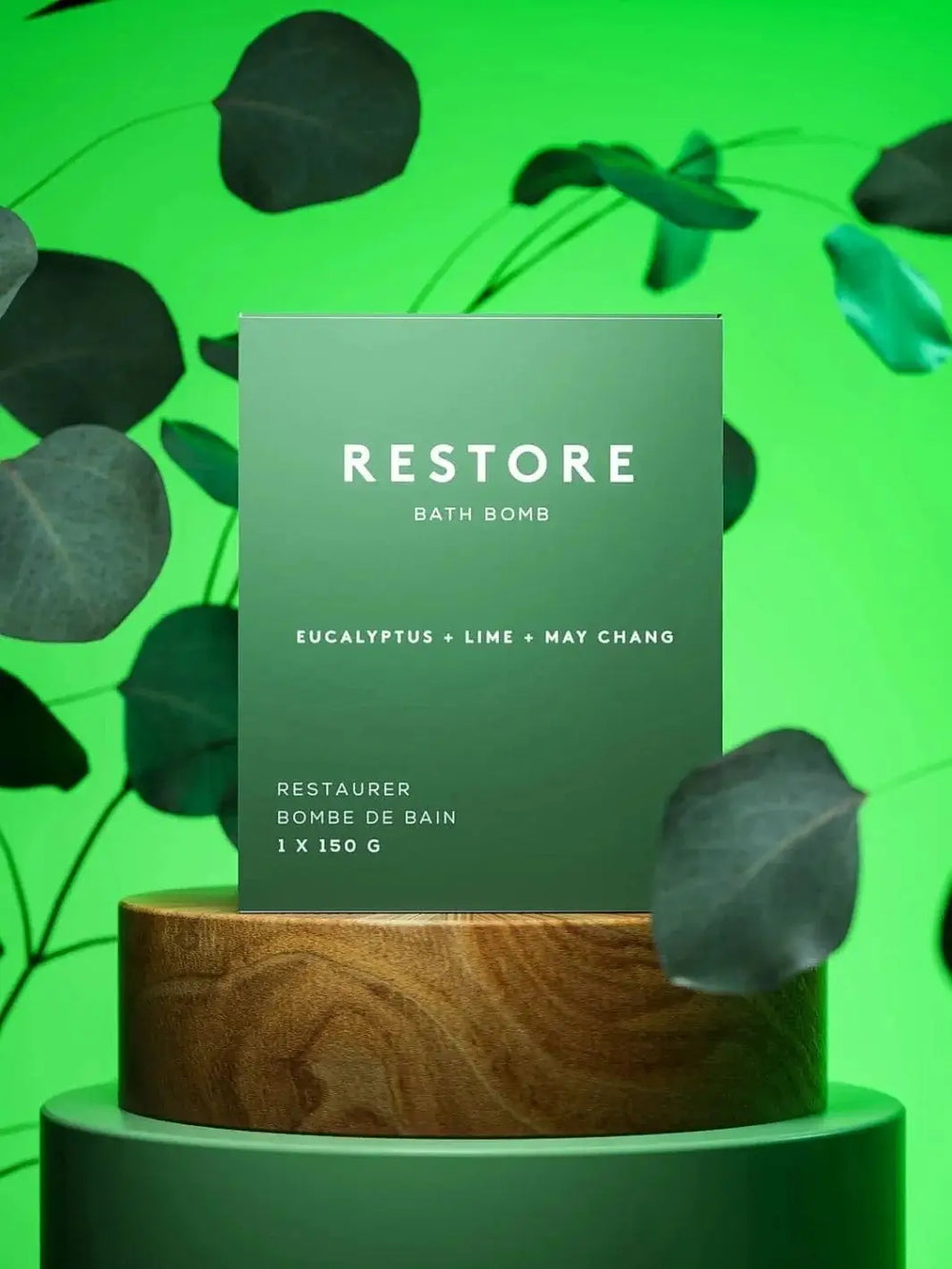 The green packaging for the Bare Skin Bar RESTORE Bath Bomb on a wooden cylindrical pedestal surrounded by eucalyptus leaves against a bright green background | Twentyseven Toronto