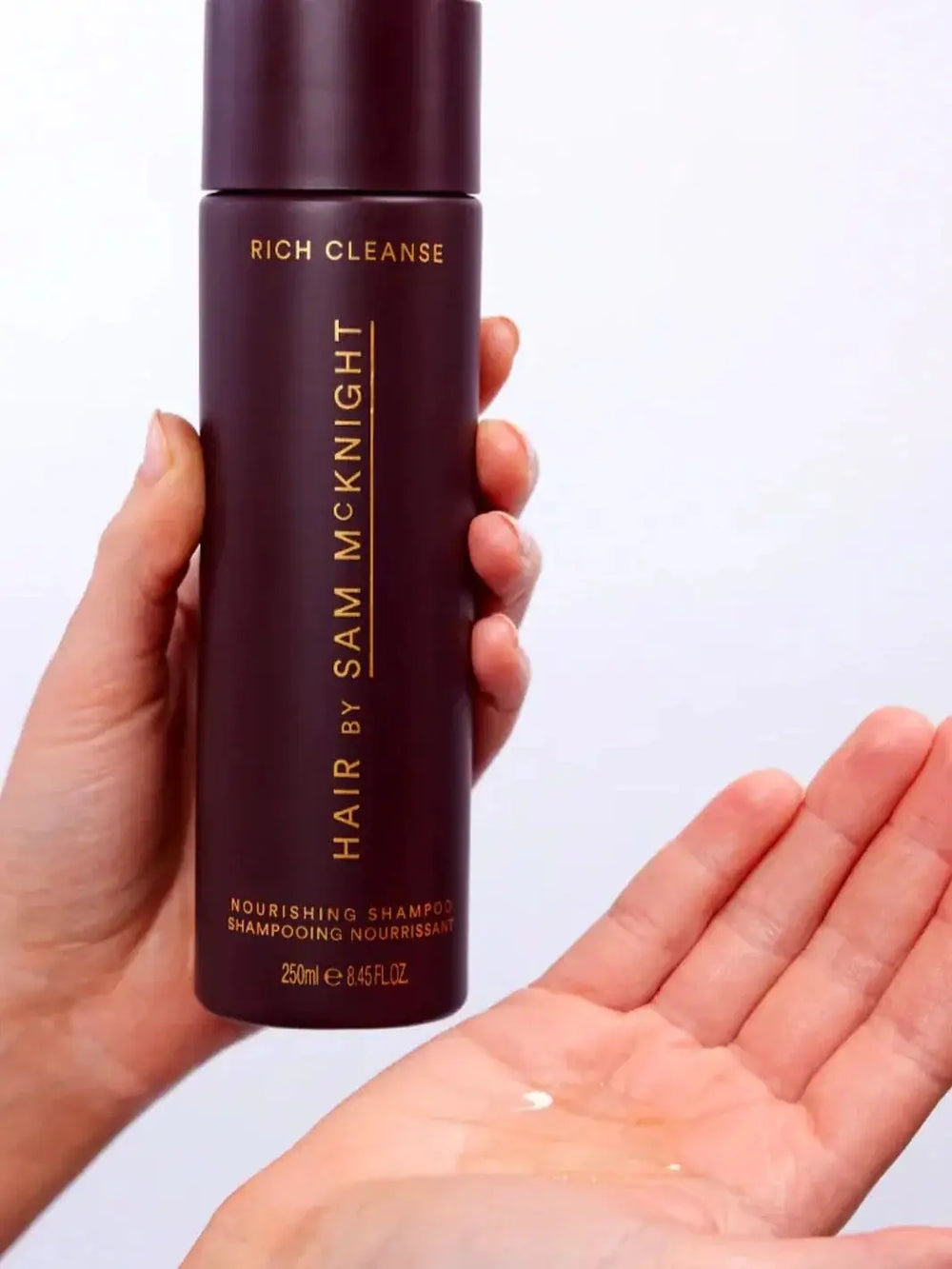 A hand holds a tall, dark purple bottle to pour a clear, liquid formula into an open palm, demonstrating the texture of Hair by Sam McKnight Rich Cleanse Nourishing Shampoo | Twentyseven Toronto