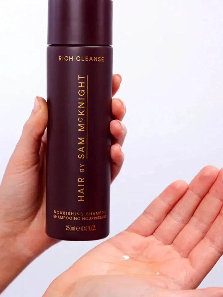 A hand holds a tall, dark purple bottle to pour a clear, liquid formula into an open palm, demonstrating the texture of Hair by Sam McKnight Rich Cleanse Nourishing Shampoo | Twentyseven Toronto