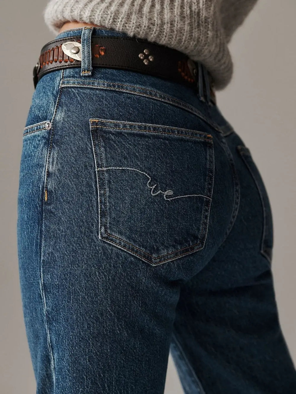 A close-up view from behind highlights the signature stitched logo embroidery on the back pocket and a dark studded leather belt loops through the Free People Risk Taker Mid-Rise Jeans - Be Bold | Twentyseven Toronto