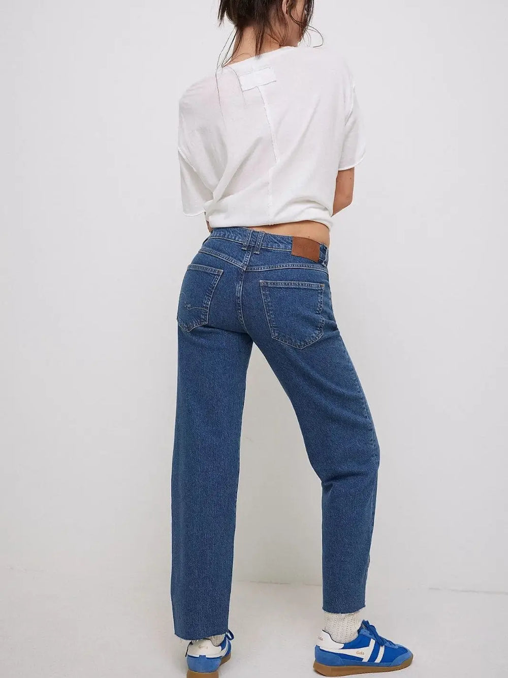 A full-length back view showcases the raw hems, relaxed fit, and leather logo patch on the waistband of the Free People Risk Taker Mid-Rise Jeans - Be Bold paired with a white tee and blue sneakers |  Twentyseven Toronto