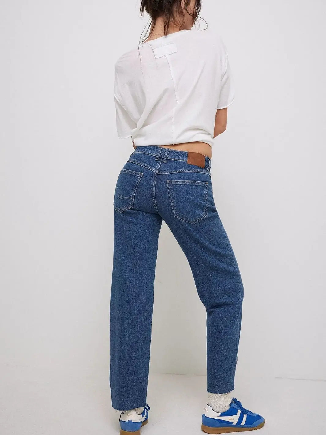 A full-length back view showcases the raw hems, relaxed fit, and leather logo patch on the waistband of the Free People Risk Taker Mid-Rise Jeans - Be Bold paired with a white tee and blue sneakers |  Twentyseven Toronto