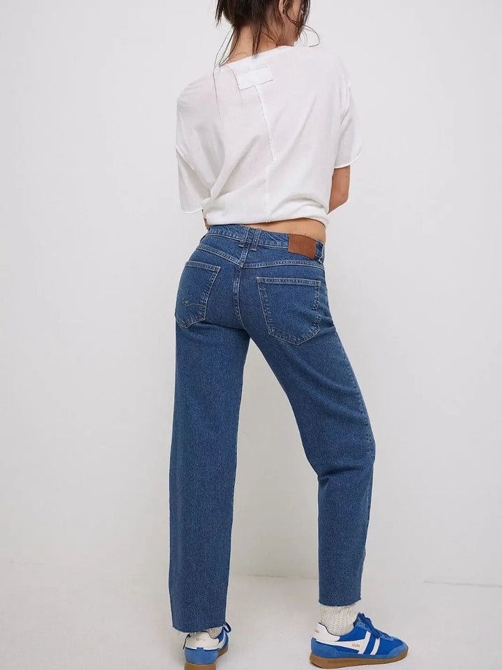 A full-length back view showcases the raw hems, relaxed fit, and leather logo patch on the waistband of the Free People Risk Taker Mid-Rise Jeans - Be Bold paired with a white tee and blue sneakers |  Twentyseven Toronto