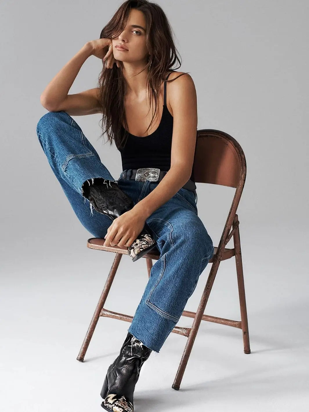 A model sits on a folding chair to display the distinct paneled leg seams and raw edge hems of the Free People Risk Taker Mid-Rise Jeans - Be Bold styled with a simple black tank top | Twentyseven Toronto