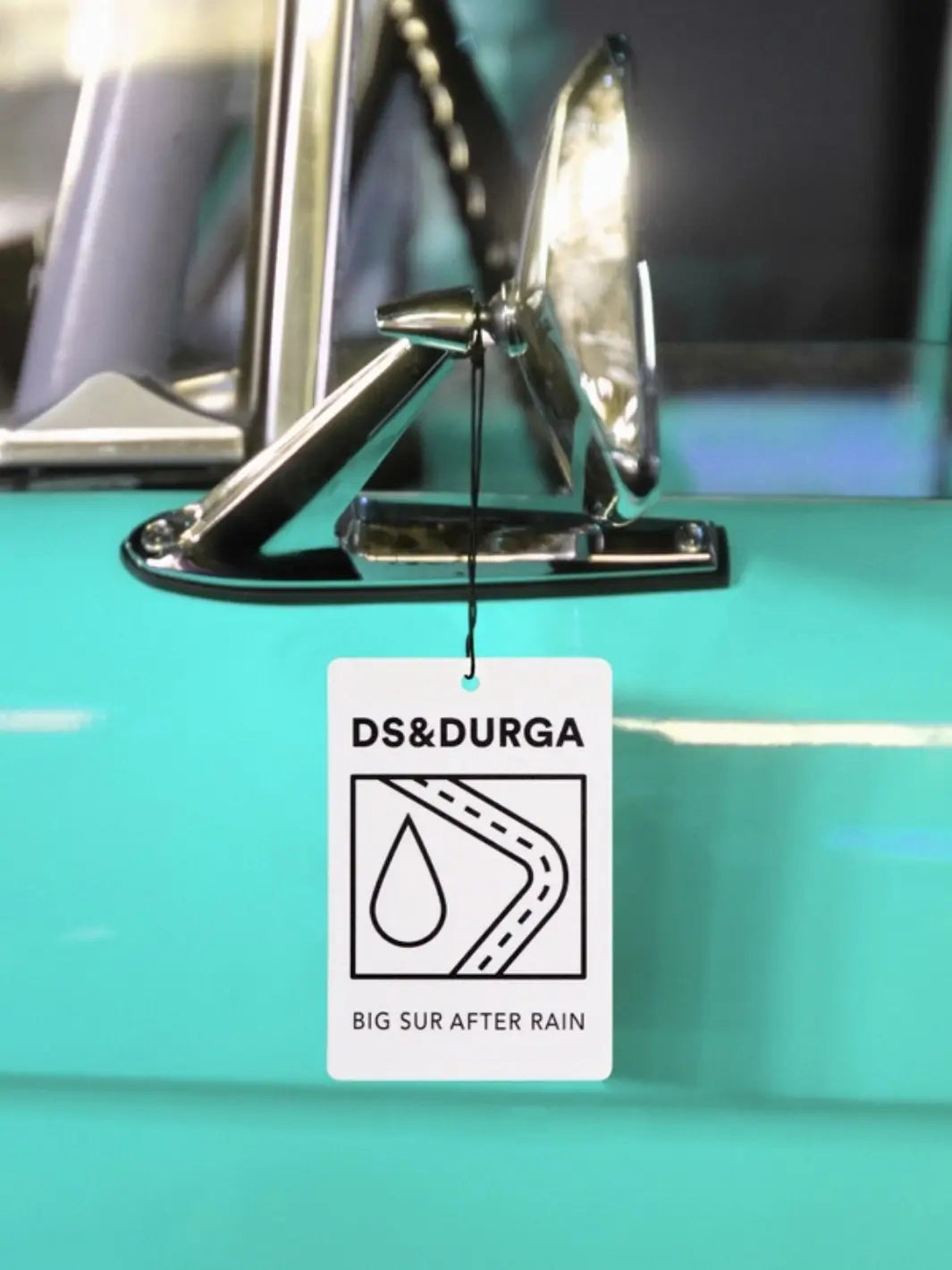 DS&Durga car air freshener 'Big Sur After Rain' hanging from a car's rearview mirror with a teal car background | Twentyseven Toronto