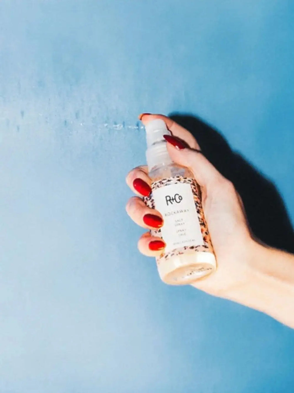 A hand with red fingernails pumps a fine mist from a bottle of R+Co ROCKAWAY Salt Spray against a solid blue backdrop | Twentyseven Toronto