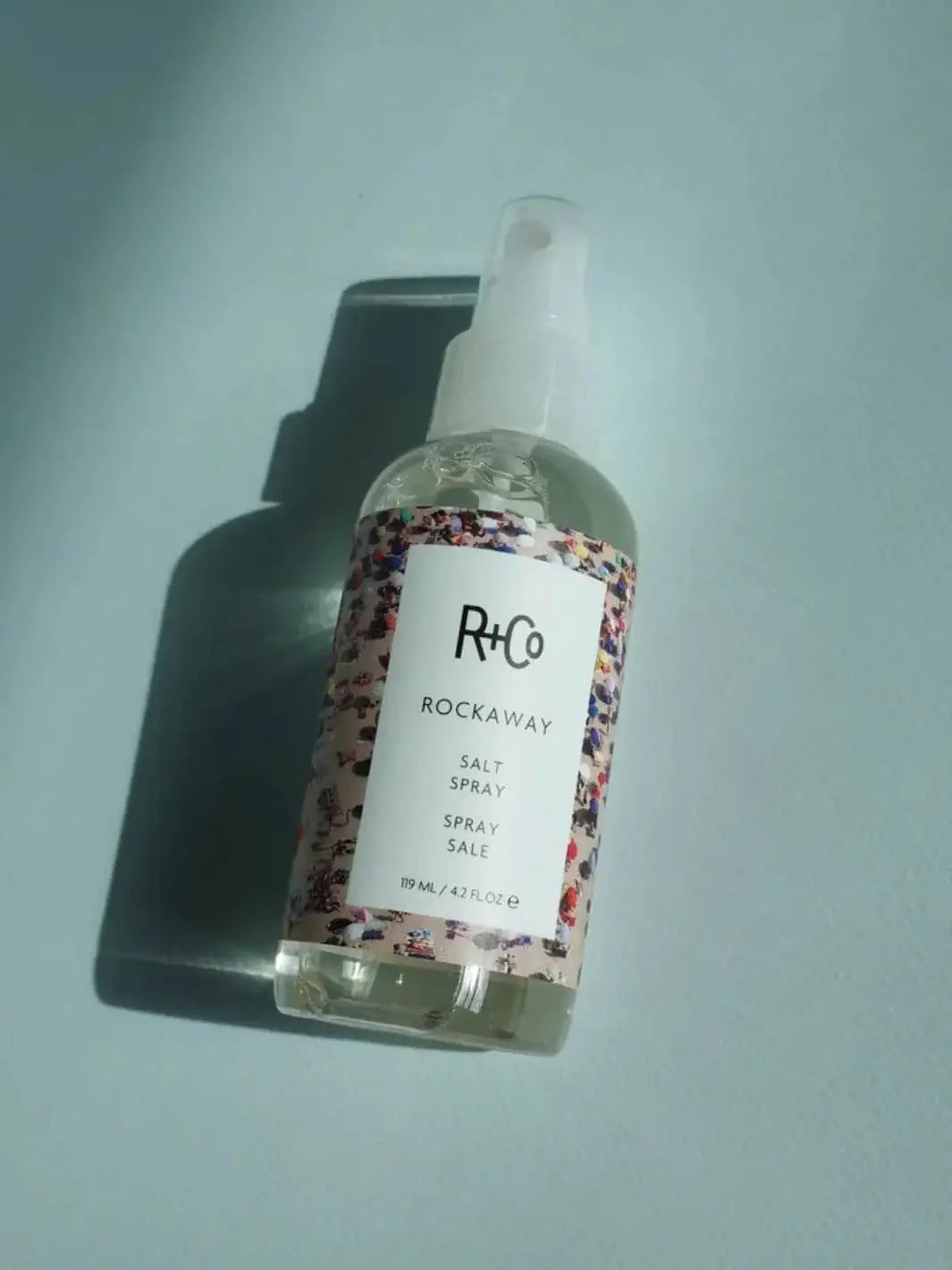 A clear spray bottle of R+Co ROCKAWAY Salt Spray rests flat on a light blue surface under bright, casting sunlight | Twentyseven Toronto