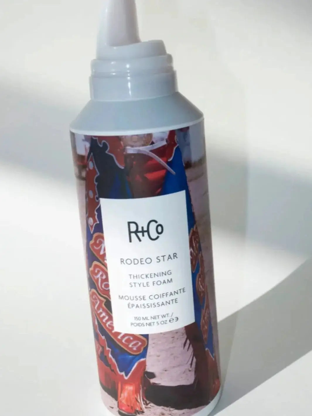 A bottle of R+Co RODEO STAR Thickening Style Foam stands with a shadow on a white background | Twentyseven Toronto