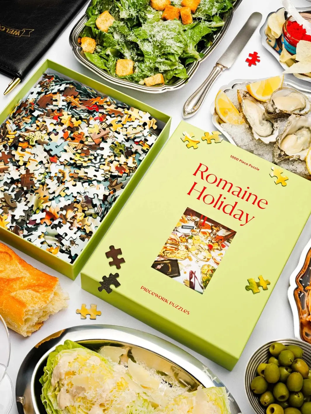 The open box of the Piecework Puzzles Romaine Holiday 1000 Piece Puzzle, filled with loose puzzle pieces, is styled on a white surface alongside fresh salads, oysters on ice, and olives | Twentyseven Toronto