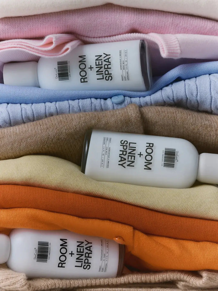 Three 100ml bottles tucked horizontally inside a colorful stack of folded sweaters, with the bottom bottle displaying the label for Dedcool Room + Linen Spray 01 "Taunt" | Twentyseven Toronto