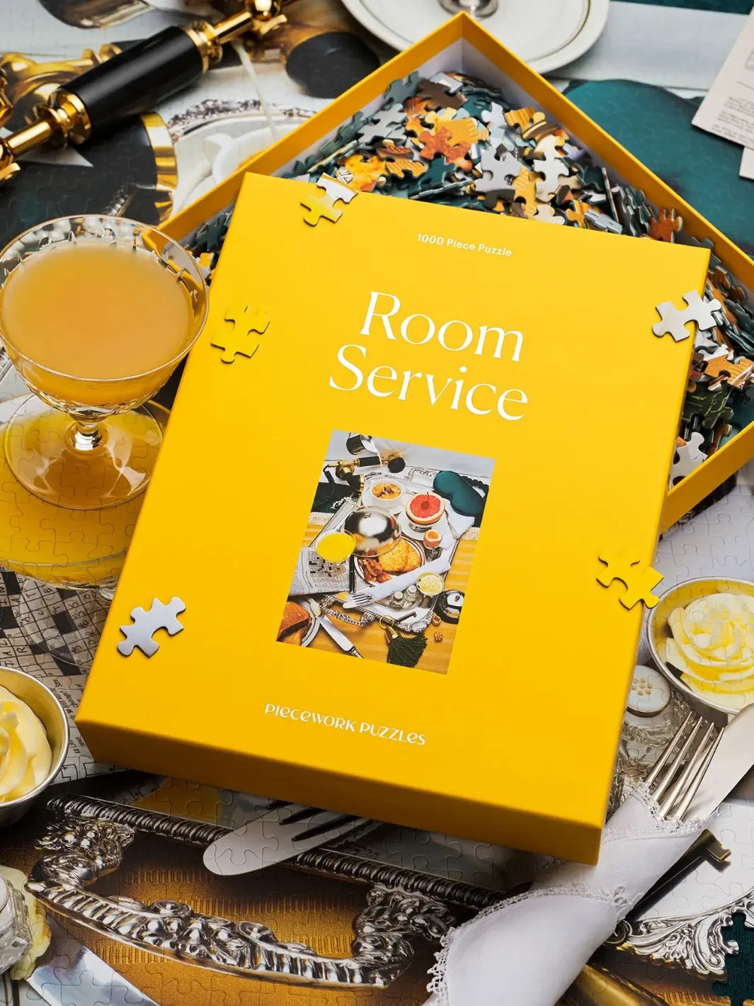 The vibrant yellow box of the Piecework Puzzles Room Service 1000 Piece Puzzle is displayed on top of a partially completed jigsaw puzzle next to a glass of orange juice | Twentyseven Toronto