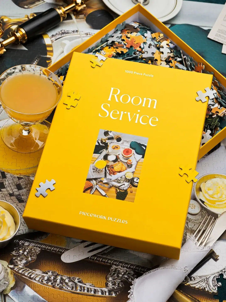 The vibrant yellow box of the Piecework Puzzles Room Service 1000 Piece Puzzle is displayed on top of a partially completed jigsaw puzzle next to a glass of orange juice | Twentyseven Toronto