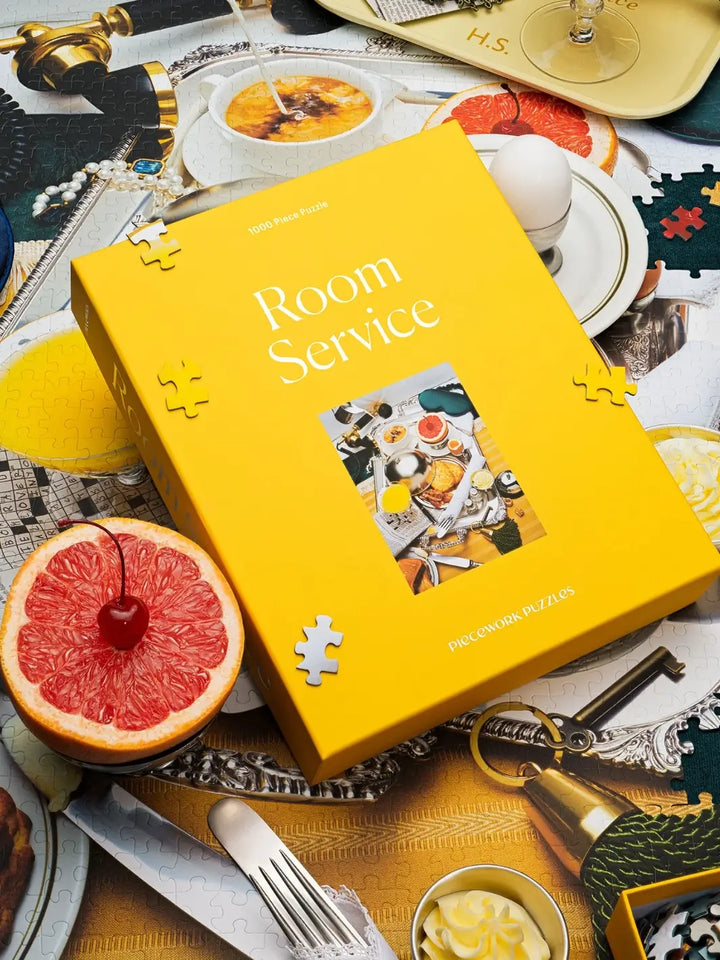 The yellow box of the Piecework Puzzles Room Service 1000 Piece Puzzle sits atop an actively assembled puzzle alongside a fresh grapefruit slice, a soft-boiled egg, and a silver fork | Twentyseven Toronto