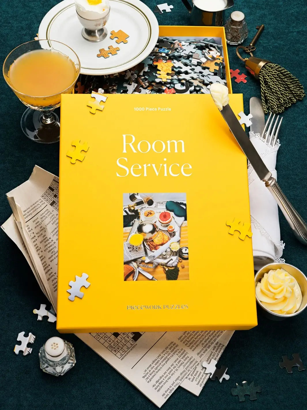 An open box of the Piecework Puzzles Room Service 1000 Piece Puzzle revealing loose puzzle pieces is creatively styled with newspaper, a butter knife, a silver fork, and a dish of whipped butter | Twentyseven Toronto