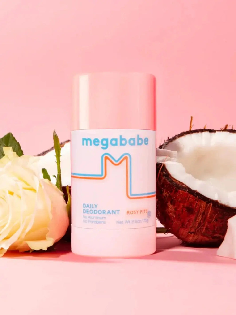A stick of Megababe Rosy Pits daily deodorant stands vertically on a pink surface flanked by a pale yellow rose and a cracked coconut half to evoke its fresh scent | Twentyseven Toronto