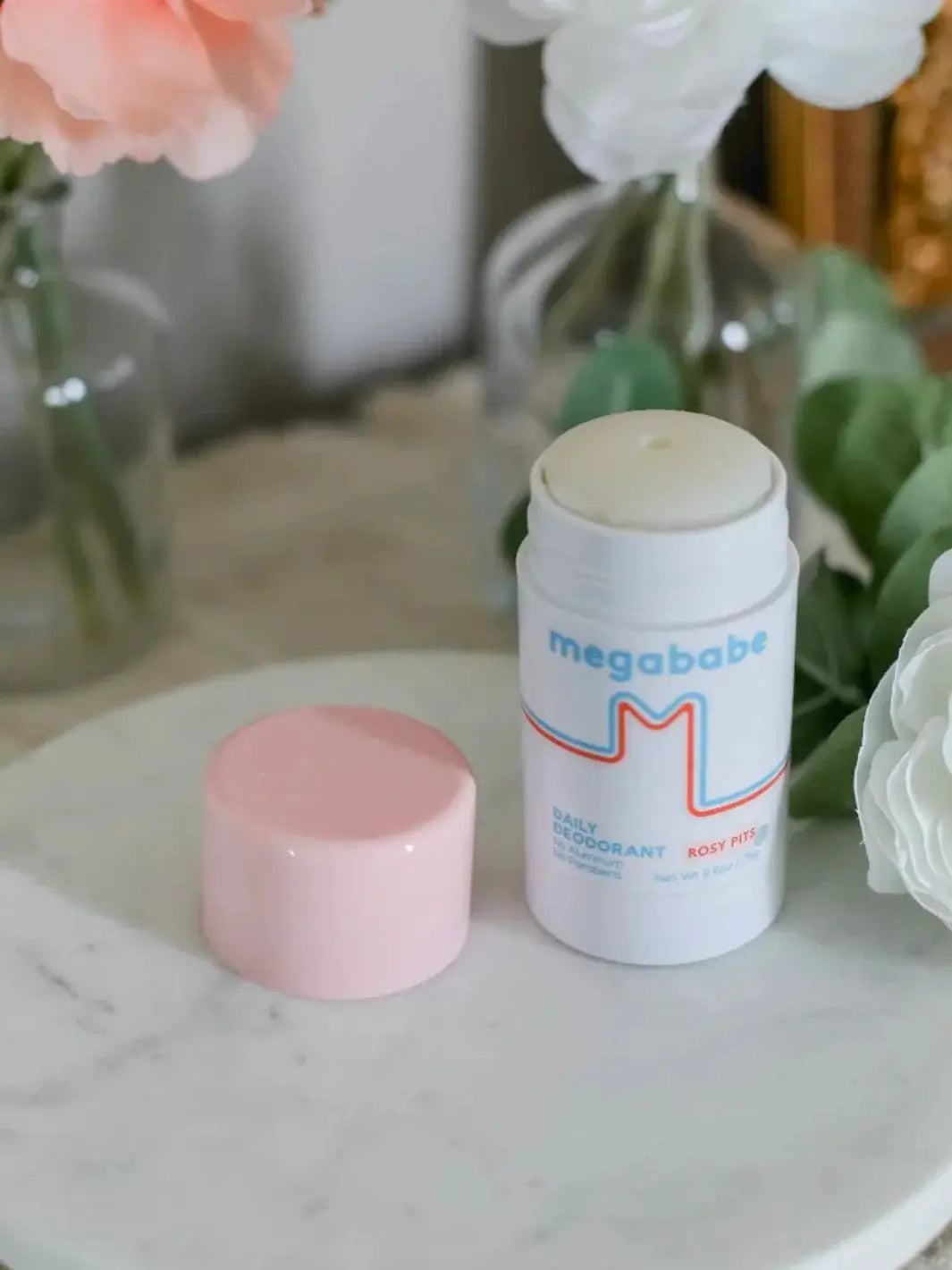 An opened container of Megababe Rosy Pits deodorant sits next to its pink cap on a white marble vanity top surrounded by delicate white roses | Twentyseven Toronto